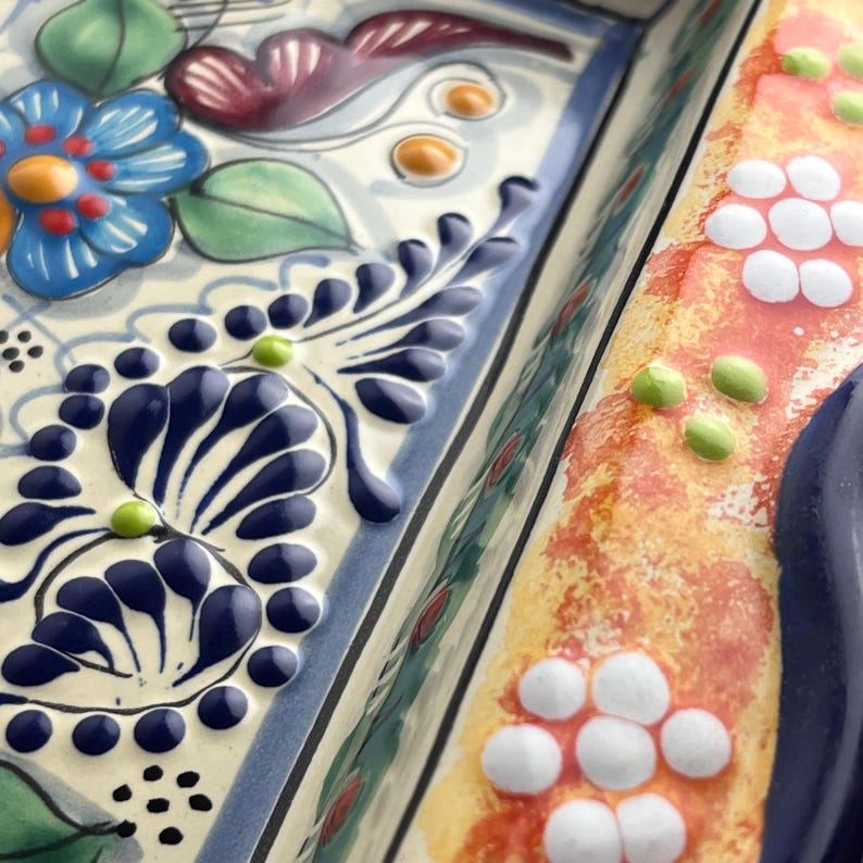 May include: Close-up of a decorative ceramic serving dish with a raised floral design. The dish features a blue flower, dark blue leaves, and white and orange accents. The edges are outlined in blue.