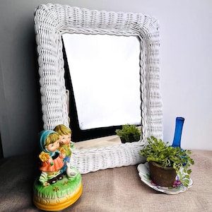 May include: A white wicker framed mirror with a rectangular shape. A ceramic figurine of two children holding flowers sits in front of the mirror. A small potted plant and a blue glass bottle are also in the image.