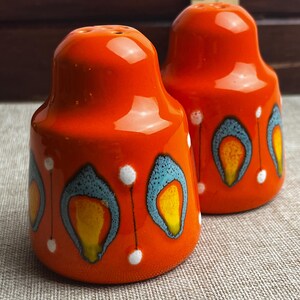 May include: A pair of orange ceramic salt and pepper shakers with a glossy finish. Each shaker has a decorative pattern of blue, yellow, and white accents. The shakers are rounded with a small hole at the top.