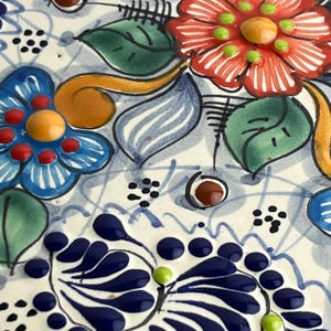 May include: Close-up of a ceramic piece with a floral design. The piece features red, blue, and orange flowers with green leaves and accents. The background is white with blue lines and dots.