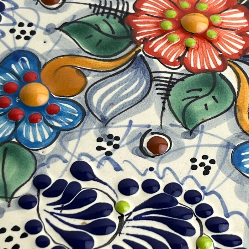May include: Close-up of a ceramic piece with a floral design. The piece features red, blue, and orange flowers with green leaves and accents. The background is white with blue lines and dots.