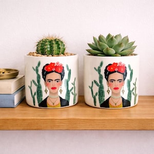 May include: Two white ceramic plant pots featuring a Frida Kahlo portrait and cactus design. One pot contains a green cactus, the other a green succulent. The pots sit on a wooden shelf, ideal for home decor.