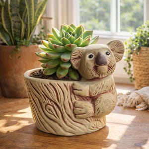 May include: A koala-shaped plant pot with a succulent. The beige ceramic pot features a tree trunk design and a koala bear figure. The succulent has green leaves with red edges. The pot is on a wooden surface.