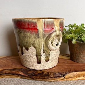 May include: A ceramic planter with a unique design. The planter has a speckled beige base, with a red and tan upper section. The glaze has a dripped effect, with tan and brown colours. A decorative handle is on the side. A small plant is in the background.