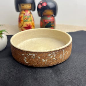 Peut inclure: Un petit plat rond en céramique avec un intérieur brun clair et un extérieur brun plus foncé. L'extérieur présente des caractères japonais blancs. Deux poupées japonaises décoratives sont en arrière-plan.