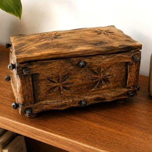 May include: A rectangular wooden box with a dark brown finish and a rough, textured surface. The lid and front panel feature carved star-shaped patterns. Dark metal studs are embedded along the edges and corners of the box structure.