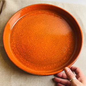 May include: A round, orange ceramic plate with a textured surface. The plate has a slightly raised rim and a glossy finish. The plate is held by a hand with a silver ring.
