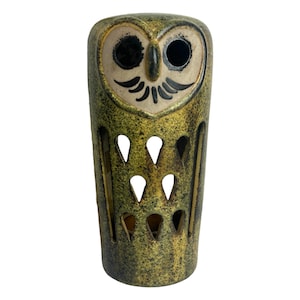 May include: A ceramic owl-shaped candle holder with a textured, mottled green and brown finish. The owl has large, dark eye sockets, a cream-colored face, and black details. The body features teardrop-shaped cutouts.