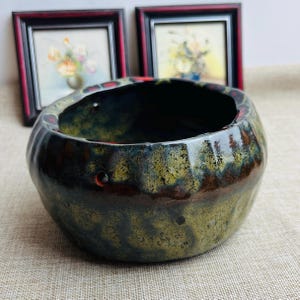 May include: A ceramic bowl with a dark, textured exterior. The bowl has a rounded shape with a wide opening and a dark interior. The exterior features a blend of green, brown, and black hues, with a glossy finish. Two small holes are visible on the side.