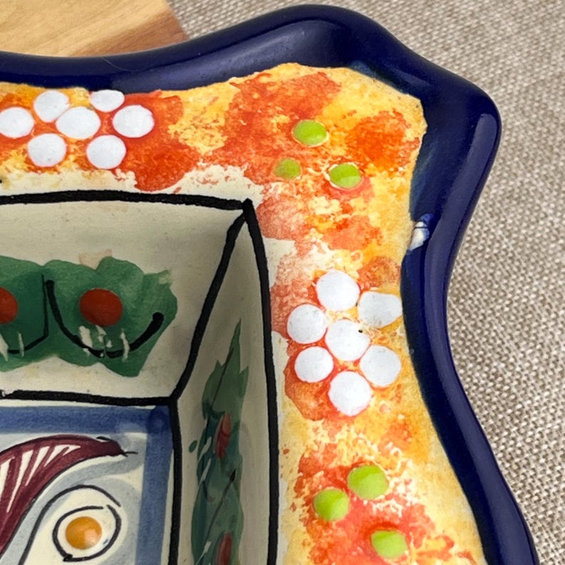 May include: A close-up of a ceramic dish with a dark blue rim and a colorful floral design. The dish features a white interior with green and red accents, and the rim is decorated with orange, yellow, and white floral patterns.