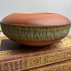 May include: A terracotta-coloured ceramic bowl with a wide, shallow design. The bowl has a band of green and brown textured glaze around the middle, and a gold rim. The bowl is a decorative piece.
