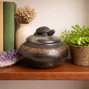 May include: A dark brown, rounded ceramic lidded jar featuring a sculpted turtle figurine on the lid. The jar has a smooth, matte finish with a horizontal band of etched, lighter brown geometric patterns encircling the centre of the vessel.
