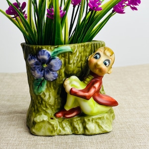 May include: A vintage ceramic planter shaped like a tree trunk, with a seated elf figurine. The planter is green with a blue flower and holds purple and green artificial flowers. The elf wears a red jacket and yellow trousers.