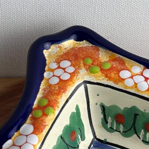 May include: A close-up of a decorative ceramic dish with a dark blue rim. The dish features a colorful design with orange, white, green, and red accents. The interior has a cream-colored background with green and red floral patterns.
