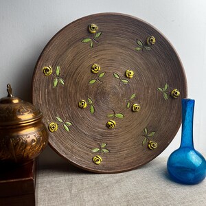 Peut inclure: Grande assiette en céramique marron avec une surface texturée et un motif floral. L'assiette présente des roses jaunes et des feuilles vertes disposées en cercle. Un petit récipient en laiton orné et un vase en verre bleu sont également visibles.