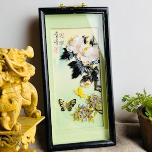 May include: Framed artwork with a floral and butterfly design. The black framed artwork has a light green background. White flowers with yellow centers and black and yellow butterflies are depicted. The artwork also has Chinese characters.