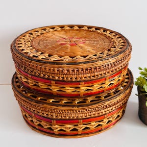 Peut inclure: Deux paniers ronds tressés empilés. Les paniers sont en osier de couleur naturelle avec des accents rouges et noirs. Les couvercles ont un motif tissé décoratif. Les paniers mesurent environ 25 cm de diamètre.