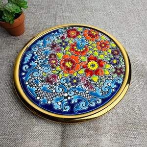 May include: A circular ceramic plate with a gold edge. The plate is decorated with a colourful floral pattern in red, orange, yellow, purple, and blue. The background is a dark blue with white filigree details.