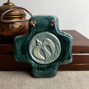 May include: A teal ceramic cross-shaped wall hanging with a raised bird design in the centre. The cross has a glossy finish and a leather loop for hanging. A copper container is visible in the background.