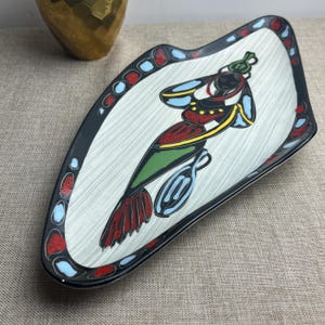 May include: A ceramic serving platter with a black rim and a colourful abstract design. The platter features a central image of a stylised figure with red, green, yellow, and blue accents. The border has red and blue circles.