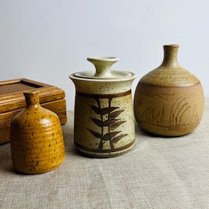 May include: A selection of three ceramic vases and a wooden box. The vases are in shades of brown and tan, with speckled and textured surfaces. One vase has a lid, another has a leaf design, and the third has an etched pattern.