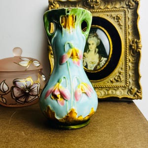 May include: A vintage ceramic vase in a pale blue colour, decorated with pink and yellow floral patterns. The vase has a distinctive shape with two side openings and a detailed, textured surface. A decorative framed portrait and a pink glass jar are in the background.