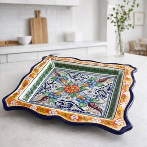 May include: A square, hand-painted ceramic serving platter with a scalloped edge. The platter features a vibrant floral design in blue, orange, green, and white against a blue background. The rim is decorated with a repeating pattern of white flowers and orange accents.