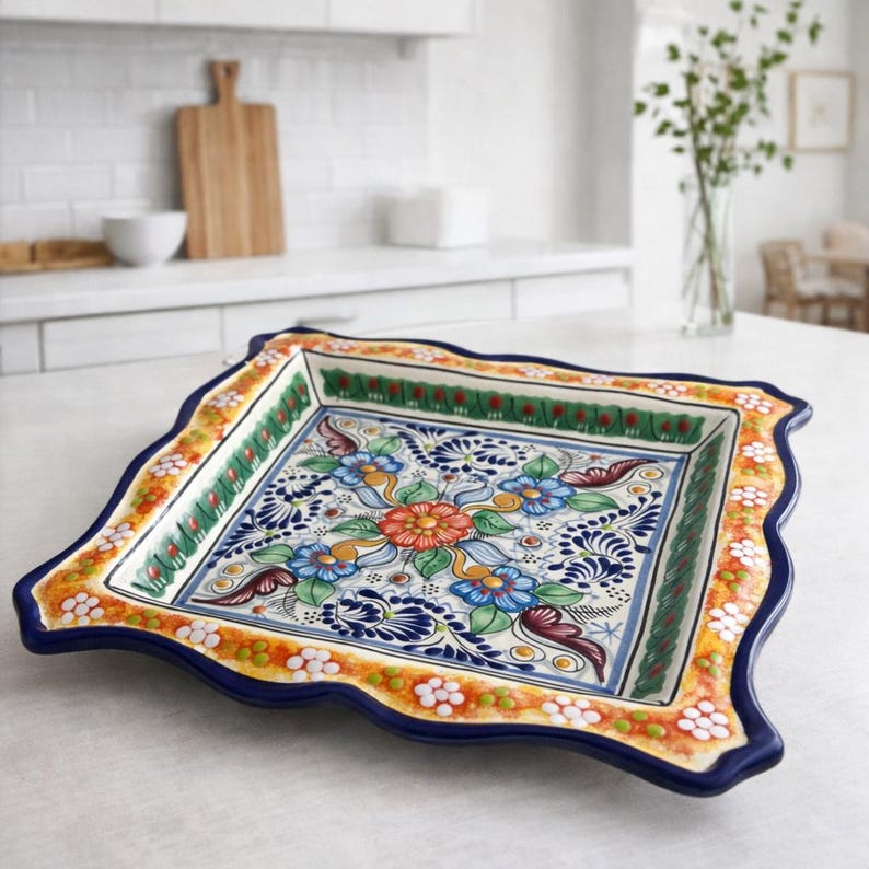 May include: A square, hand-painted ceramic serving platter with a scalloped edge. The platter features a vibrant floral design in blue, orange, green, and white against a blue background. The rim is decorated with a repeating pattern of white flowers and orange accents.