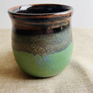 May include: A handmade ceramic tumbler with a rounded base. The top rim is dark brown, transitioning to black, then a speckled gray-brown, and finally a vibrant green. The interior is dark brown.