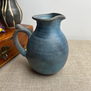 May include: A handmade, blue ceramic jug with a rounded body and a curved handle. The jug has a ruffled rim and a textured surface. The colour transitions from a lighter blue at the top to a darker blue at the base. A decorative piece for the home.