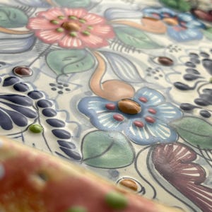 May include: Close-up of a ceramic tray with a floral design. The tray features pink, blue, and purple flowers with green leaves and brown accents. The background is a cream color with a subtle pattern.