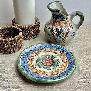 May include: A small, white ceramic plate with a green, blue and orange floral design. The plate is decorated with a large, pink flower in the centre.