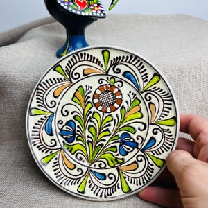 May include: A small, round, hand-painted ceramic plate with a floral design. The plate has a cream-coloured background with a central flower and surrounding leaves in green, blue, orange, and brown. Black outlines define the design.