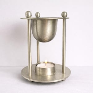 May include: A silver aromatherapy oil warmer with a lit tea light candle. The warmer has a round base, vertical supports, and a bowl-shaped top. Three small spheres are on top of the warmer.