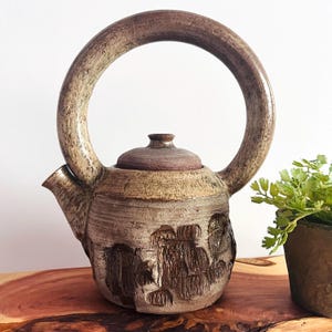 May include: A handmade ceramic teapot with a large, arched handle. The teapot is mainly brown with textured details and a lid. The body features a carved design. The teapot is displayed on a wooden surface.