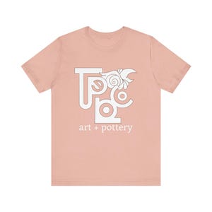 May include: A light pink t-shirt featuring a white graphic design of the letters "TPC" with a stylised floral design. The text "art + pottery" is printed below the graphic.