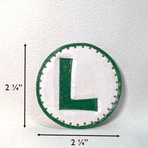 May include: A round, white and green decorative item with the letter "L" in green. The item is 2 1/4 inches in diameter and 2 1/4 inches in height. The border is green with small brown dots. The background is white.