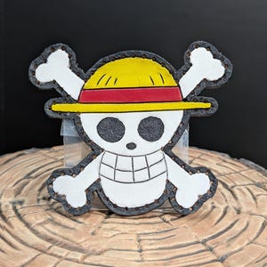 May include: A white skull and crossbones with a yellow and red straw hat on top. The skull and crossbones are outlined in black and have a stitched appearance.