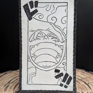 May include: A white leather patch with black stitching and a black and white illustration of a character's face with a large open mouth. The character's face is surrounded by a black frame with the Japanese characters "ギ" and "ガ" on the top and bottom.