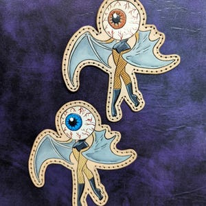 May include: Two whimsical, die-cut patches featuring an eyeball with bat wings, fishnet stockings, and boots. One has a brown iris, the other a blue iris. The patches are outlined with a dotted border and set against a purple background.