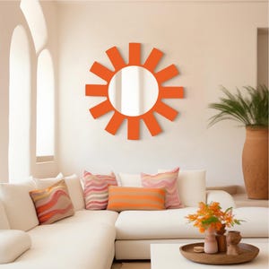Puede incluir: Un gran espejo de sol naranja con un centro reflectante redondo cuelga de una pared blanca sobre un sofá blanco con almohadas naranjas y rosas.