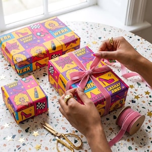 May include: Gift boxes with colorful, patterned wrapping paper featuring a drag queen illustration, high heels, and lipstick. The boxes are tied with pink velvet ribbon. The text on the boxes reads "WERK", "SASHAY AWAY", and "SHANTAY YOU STAY".