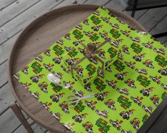 May include: A wrapped gift box and wrapping paper with a repeating pattern of monster trucks and the text "Get the Ghoul Times Roll" on a bright green background. A brown ribbon and scissors are also visible.