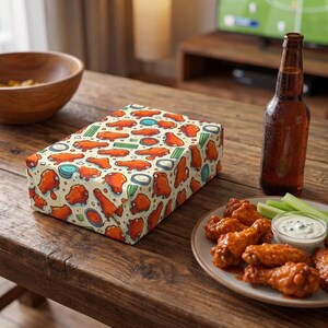 May include: A rectangular gift box with a repeating pattern of orange chicken wings, blue bowls, and green celery on a cream background. A plate of chicken wings, celery, and dip, and a beer bottle are on a wooden table.
