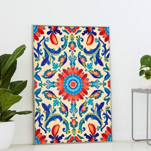 Puede incluir: Una impresión artística enmarcada con un diseño floral simétrico en rojo, azul y amarillo. La obra de arte tiene una flor central con elementos florales y de desplazamiento circundantes. La impresión se muestra contra una pared blanca, con plantas en primer plano.