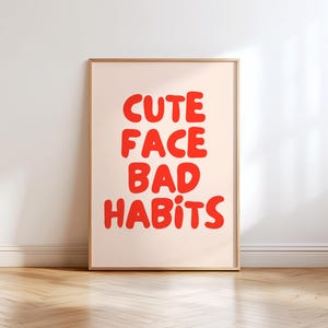 Pode incluir: Uma impressão artística emoldurada com as palavras "CUTE FACE BAD HABITS" em letras vermelhas em negrito. O texto está centrado em um fundo pêssego claro. A moldura é de madeira natural fina. A impressão é exibida contra uma parede branca.
