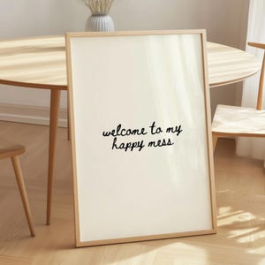 Pode incluir: Uma impressão emoldurada com o texto "welcome to my happy mess" em escrita preta. A impressão está encostada a uma parede, com uma mesa e uma cadeira de madeira no fundo. O quarto tem uma paleta de cores neutras.