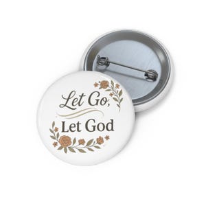 May include: White button pin with the text "Let Go, Let God" in a script font, surrounded by floral accents in shades of brown and green. The pin has a metal backing and a safety pin closure.