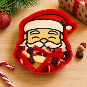 May include: A red silicone tray shaped like a Santa Claus face, filled with assorted gummy candies including red licorice strips, dark berry-shaped gummies, and cream-colored pieces. The tray features a printed graphic of a smiling Santa.