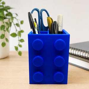 May include: A bright blue rectangular desk organiser shaped like a building block with six circular studs on the front. The container is divided into two sections and holds various office supplies including pens, a marker, and blue scissors.